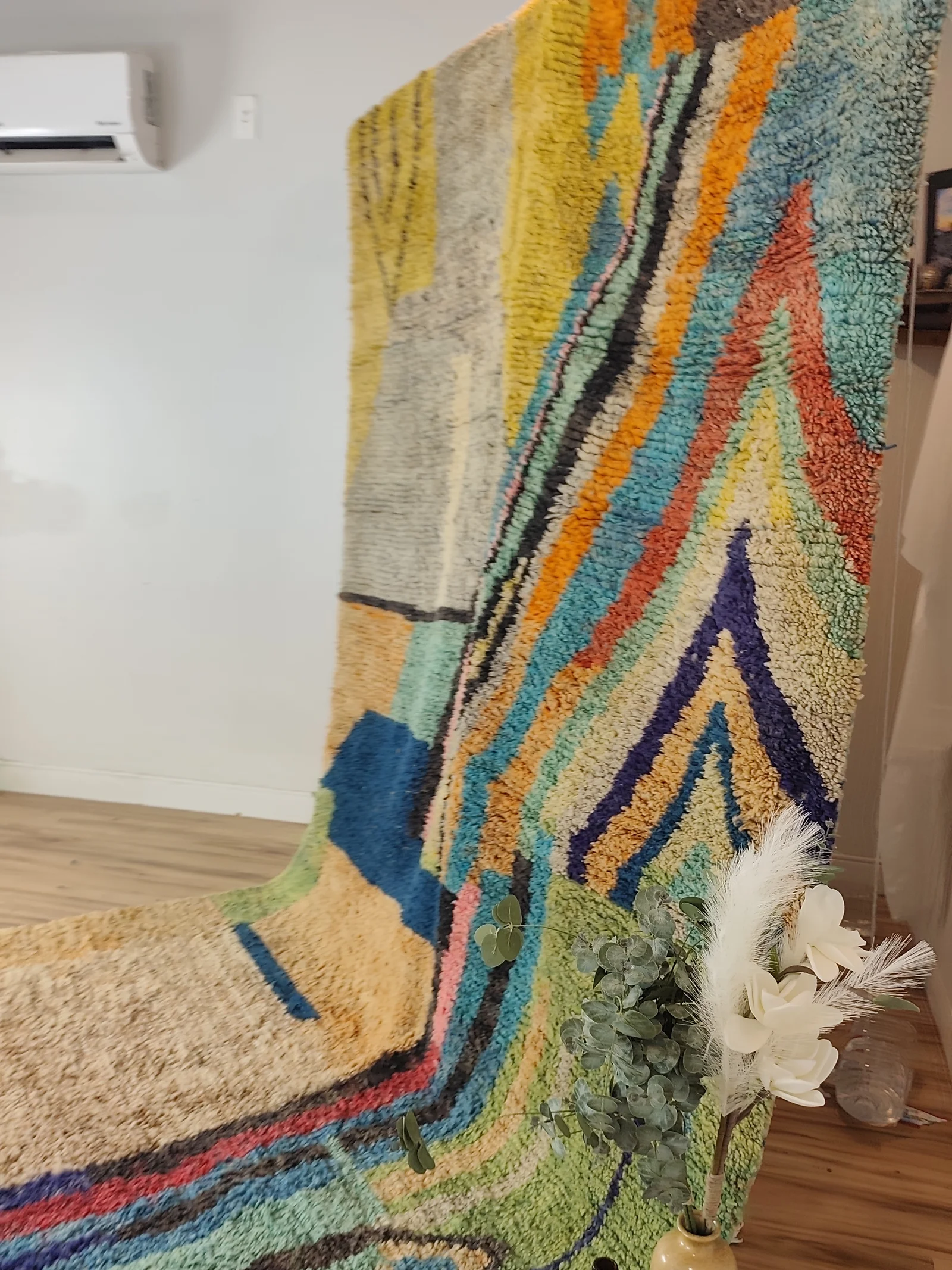 Soukaina — the hung rug falling to the floor beside eucalyptus, an abstract Boujaad in teal, blush and mustard, with bold diagonal bands cutting across a cream ground, 323 × 178 cm, view 13 of 20